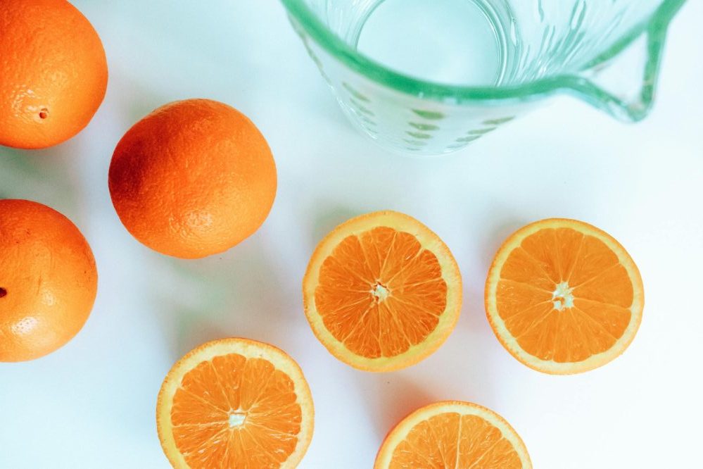 A vibrant still life of whole and sliced oranges next to a glass pitcher.